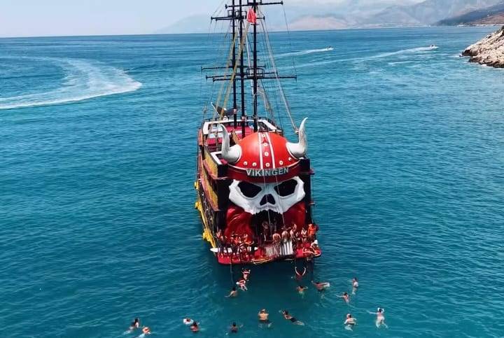 The Vikingen, the red Viking-style boat, at anchor in turquoise water off Saranda, seen from a drone, with swimmers around it
