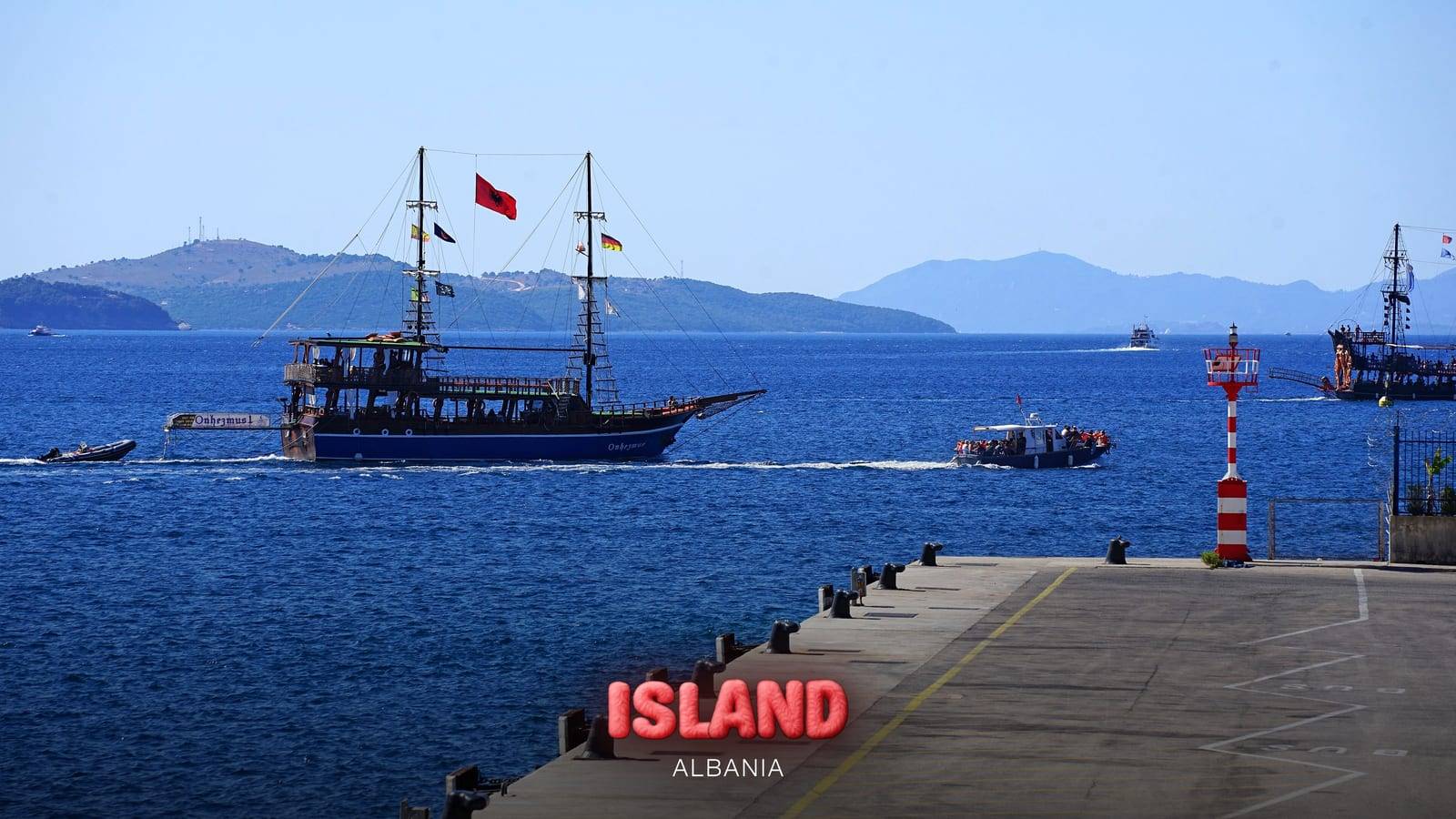 A two-masted excursion boat leaving the port of Saranda with Corfu behind, Albania, with the Party Island Albania logo