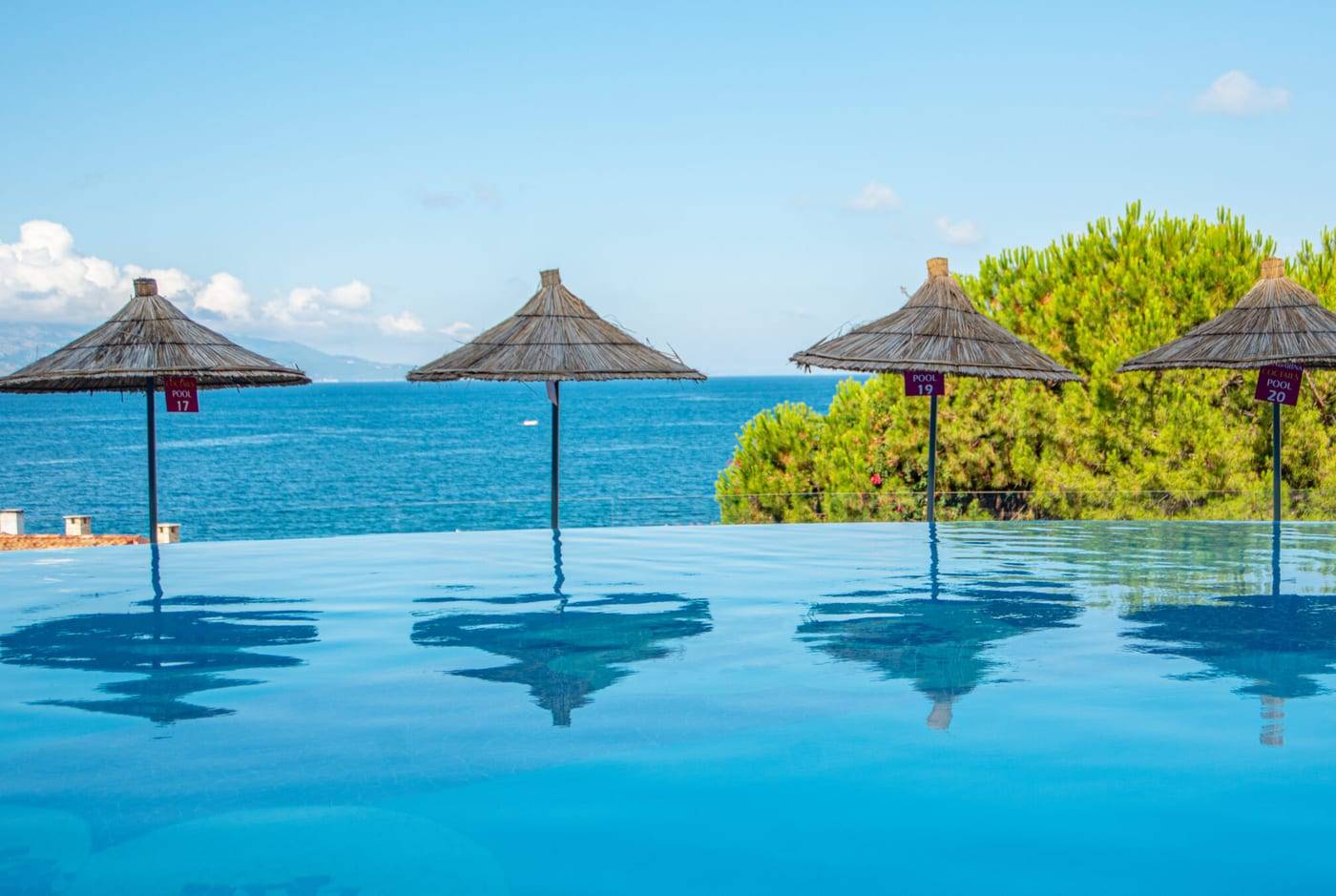 The infinity pool at Bougainville Bay Resort & SPA in Saranda, with straw parasols reflected in the water and the sea beyond