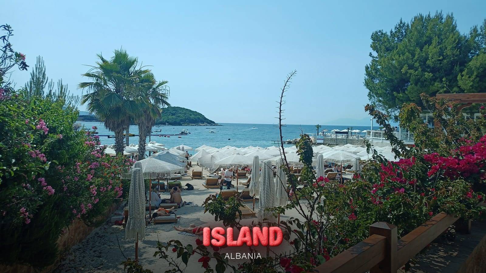 Ksamil beach seen between bougainvillea and palm trees, with parasols and a wooded islet offshore, Albania, with the Party Island Albania logo